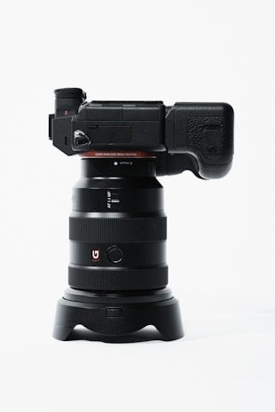 A professional digital camera with a large lens is positioned vertically. The camera body is black with a textured grip and various controls on the side. The lens has a wide barrel with multiple rings and a red 'G' logo. The setup is placed on a minimalistic white background, emphasizing the sleek design of the camera.