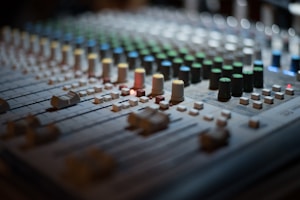A close-up view of a professional audio mixing console featuring numerous knobs and sliders in various colors. The focus is on the details of the controls, with some lit up indicators providing visual feedback. The image captures the complexity and precision involved in audio mixing.