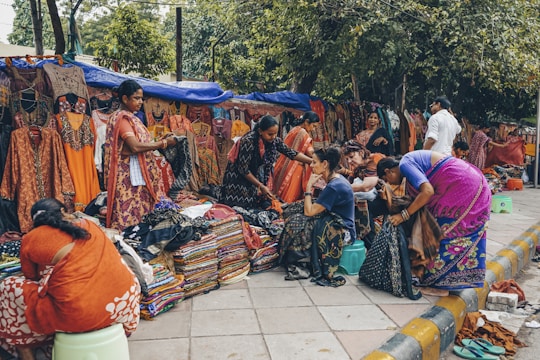 A bustling outdoor market scene with several women browsing and selling colorful traditional clothing. The garments, including vibrant sarees and dresses, are displayed both hanging and stacked on the sidewalk. The setting is lively with people interacting, some seated on small stools while others stand or browse through the fabrics. Trees and makeshift stalls form the backdrop.