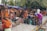 A bustling outdoor market scene with several women browsing and selling colorful traditional clothing. The garments, including vibrant sarees and dresses, are displayed both hanging and stacked on the sidewalk. The setting is lively with people interacting, some seated on small stools while others stand or browse through the fabrics. Trees and makeshift stalls form the backdrop.