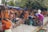 A bustling outdoor market scene with several women browsing and selling colorful traditional clothing. The garments, including vibrant sarees and dresses, are displayed both hanging and stacked on the sidewalk. The setting is lively with people interacting, some seated on small stools while others stand or browse through the fabrics. Trees and makeshift stalls form the backdrop.