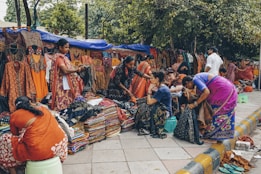 A bustling outdoor market scene with several women browsing and selling colorful traditional clothing. The garments, including vibrant sarees and dresses, are displayed both hanging and stacked on the sidewalk. The setting is lively with people interacting, some seated on small stools while others stand or browse through the fabrics. Trees and makeshift stalls form the backdrop.