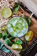 A refreshing beverage served in a glass filled with ice cubes, garnished with mint leaves. Surrounding the glass are cut lime halves and whole mint sprigs, arranged on a wooden surface with some straw decoration.