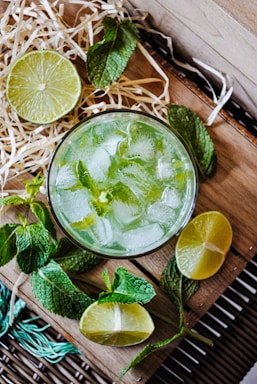 A refreshing beverage served in a glass filled with ice cubes, garnished with mint leaves. Surrounding the glass are cut lime halves and whole mint sprigs, arranged on a wooden surface with some straw decoration.