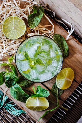 A refreshing beverage served in a glass filled with ice cubes, garnished with mint leaves. Surrounding the glass are cut lime halves and whole mint sprigs, arranged on a wooden surface with some straw decoration.
