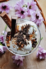 A glass filled with vanilla ice cream topped with crushed chocolate cookies and two chocolate sticks. The serving is placed on a wooden surface surrounded by light purple flowers, adding a floral touch to the presentation.