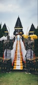 A traditional Balinese temple entrance featuring a central stone staircase draped with vibrant yellow, white, and black checkered cloths. Flanking the stairs are stone guardian statues, surrounded by multiple ornate umbrellas. The background consists of lush green trees and the sky above is overcast.