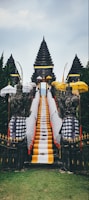 A traditional Balinese temple entrance featuring a central stone staircase draped with vibrant yellow, white, and black checkered cloths. Flanking the stairs are stone guardian statues, surrounded by multiple ornate umbrellas. The background consists of lush green trees and the sky above is overcast.