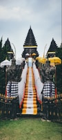 A traditional Balinese temple entrance featuring a central stone staircase draped with vibrant yellow, white, and black checkered cloths. Flanking the stairs are stone guardian statues, surrounded by multiple ornate umbrellas. The background consists of lush green trees and the sky above is overcast.