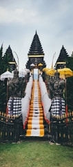 A traditional Balinese temple entrance featuring a central stone staircase draped with vibrant yellow, white, and black checkered cloths. Flanking the stairs are stone guardian statues, surrounded by multiple ornate umbrellas. The background consists of lush green trees and the sky above is overcast.