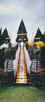 A traditional Balinese temple entrance featuring a central stone staircase draped with vibrant yellow, white, and black checkered cloths. Flanking the stairs are stone guardian statues, surrounded by multiple ornate umbrellas. The background consists of lush green trees and the sky above is overcast.