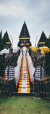A traditional Balinese temple entrance featuring a central stone staircase draped with vibrant yellow, white, and black checkered cloths. Flanking the stairs are stone guardian statues, surrounded by multiple ornate umbrellas. The background consists of lush green trees and the sky above is overcast.