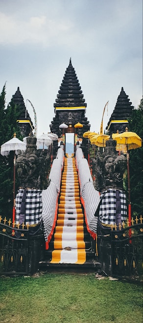 A traditional Balinese temple entrance featuring a central stone staircase draped with vibrant yellow, white, and black checkered cloths. Flanking the stairs are stone guardian statues, surrounded by multiple ornate umbrellas. The background consists of lush green trees and the sky above is overcast.