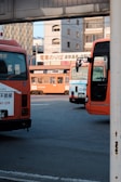 Several buses are parked at a station near an urban area. There are buildings in the background and a sign above one of the buses. The setting appears to be a bus station with multiple orange buses.