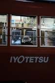 An individual wearing a hat is seated inside a tram with the brand name 'Iyotetsu' visible on its exterior. The tram has multiple windows and is illuminated from within, showcasing various handles for standing passengers. The setting appears to be urban, with some greenery visible through the windows.
