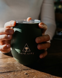 A person is holding a dark green tumbler with both hands. The tumbler features a logo with a triangle and a palm tree, along with the text 'CAYE.LIFE'. Sunlight casts soft shadows on the wooden surface beneath.