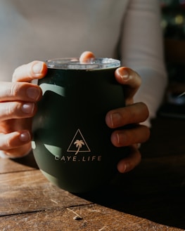 A person is holding a dark green tumbler with both hands. The tumbler features a logo with a triangle and a palm tree, along with the text 'CAYE.LIFE'. Sunlight casts soft shadows on the wooden surface beneath.