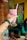 A hand with tattoos and a wristwatch holds up a tall glass of pink smoothie or shake with red syrup swirls, topped with a straw. The background includes a colorful, abstract wall mural and a person partly visible wearing a teal garment in a casual dining setting, with a table that has cups, a bottle, and a pizza.