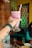 A hand with tattoos and a wristwatch holds up a tall glass of pink smoothie or shake with red syrup swirls, topped with a straw. The background includes a colorful, abstract wall mural and a person partly visible wearing a teal garment in a casual dining setting, with a table that has cups, a bottle, and a pizza.