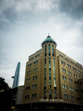 A tall, beige building with multiple windows and a distinctive glass dome at the top, displaying the words 'ARTEK SAIGON' and 'LOUIS VUITTON'. In the background, a modern skyscraper with a unique spiral design rises against a cloudy sky.