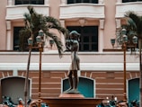 A bronze statue of a young girl playing a flute stands prominently in an urban plaza setting. The statue is surrounded by decorative street lamps and palm trees, with a classical building facade in the background. Several motorcycles are parked near the base of the statue.
