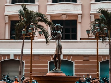 A bronze statue of a young girl playing a flute stands prominently in an urban plaza setting. The statue is surrounded by decorative street lamps and palm trees, with a classical building facade in the background. Several motorcycles are parked near the base of the statue.