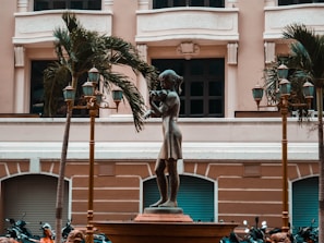 A bronze statue of a young girl playing a flute stands prominently in an urban plaza setting. The statue is surrounded by decorative street lamps and palm trees, with a classical building facade in the background. Several motorcycles are parked near the base of the statue.