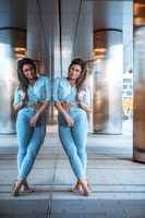 A person stands in front of a reflective surface, creating the illusion of a double. They are wearing a light blue shirt and jeans with brown sandals. The background features large metallic pillars and a tiled floor, possibly in an urban setting.