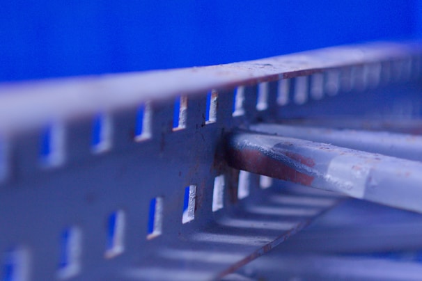 A close-up of a metal structure featuring a series of evenly spaced rectangular cut-outs. The metal appears slightly rusted in places, suggesting age or exposure to the elements. The background is a vivid blue, highlighting the industrial aesthetic.