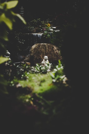 A serene garden scene featuring a stone sculpture of a meditative figure at the center. The sculpture is set against a backdrop of dark greenery and a gentle waterfall cascading over rocks. Sunlight illuminates a single yellow sunflower and some foliage in the background, creating a peaceful and tranquil atmosphere.