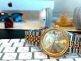 A close-up of a luxury watch with a silver and gold bracelet, featuring a gold bezel and a date display. The watch rests on a computer keyboard in an office setting, with a blurred background showing sunglasses, headphones, and a decorative pyramid.