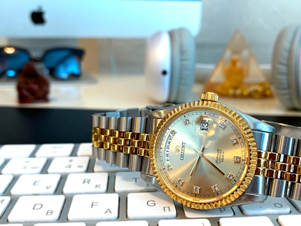 A close-up of a luxury watch with a silver and gold bracelet, featuring a gold bezel and a date display. The watch rests on a computer keyboard in an office setting, with a blurred background showing sunglasses, headphones, and a decorative pyramid.