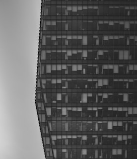 A black and white architectural photograph showcasing a large modern office building with a grid-like pattern of windows. Each floor is visible, revealing some of the interior elements such as lights and blinds.