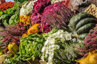 An abundant display of colorful vegetables and flowers, including a green squash, leafy greens, chrysanthemums, and heather. The vivid colors range from deep greens to vibrant reds and whites, creating a lively and bountiful harvest scene.