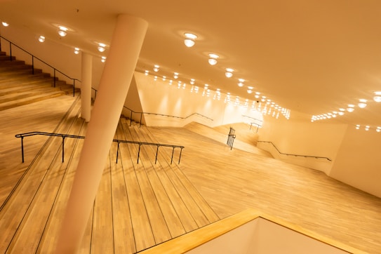 A spacious interior with a modern architectural design featuring wide wooden staircases and numerous round ceiling lights casting a warm glow. The stairs have sleek black railings, and the walls are plain with a light, neutral tone. The perspective provides a dynamic view, incorporating angles and lines that create a sense of depth and movement.