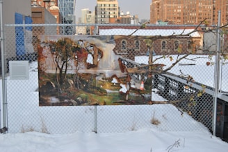 A colorful painting is mounted on a chain-link fence, depicting trees and a natural scene. The background shows an urban environment with brick buildings and skyscrapers under a cloudy sky. The ground is covered with snow, and the fence also has a small sign.