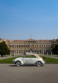 A vintage white convertible car is parked in front of an elegant, historical building with neoclassical architecture. The area is surrounded by well-maintained gardens, and the sky is clear blue. The building features numerous windows, a central fountain, and an Italian flag is visible on top.