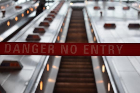 A red tape with the words 'Danger No Entry' is prominently displayed in front of an escalator. The escalator is not in operation and appears to be in a public transit or commercial area, with stainless steel sides and lighting fixtures along the edges.