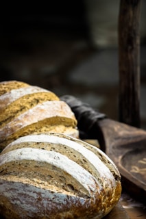 Loaves of freshly baked bread with a rustic crust are displayed on a wooden surface. The bread features a dusting of flour and a pattern of slashes on the top, giving it an artisanal appearance. In the background, there is a dark, blurred setting that enhances the focus on the bread.