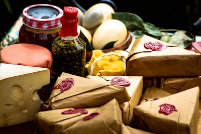 An assortment of packaged goods is displayed, including brown paper-wrapped parcels with red wax seals, a wedge of cheese with holes, a jar with a red and white checked lid, a bottle containing green peppercorns, eggs, and some leafy greens. The arrangement suggests a rustic, artisanal market setting.