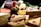 An assortment of packaged goods is displayed, including brown paper-wrapped parcels with red wax seals, a wedge of cheese with holes, a jar with a red and white checked lid, a bottle containing green peppercorns, eggs, and some leafy greens. The arrangement suggests a rustic, artisanal market setting.