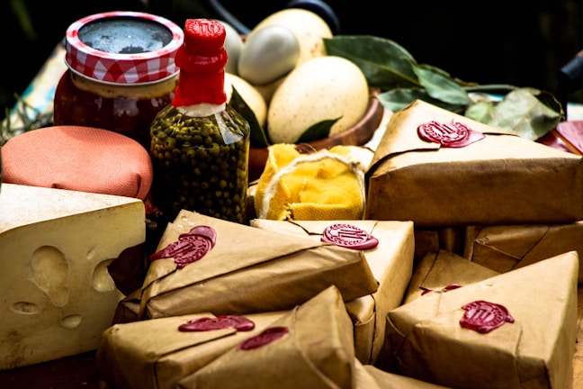 An assortment of packaged goods is displayed, including brown paper-wrapped parcels with red wax seals, a wedge of cheese with holes, a jar with a red and white checked lid, a bottle containing green peppercorns, eggs, and some leafy greens. The arrangement suggests a rustic, artisanal market setting.