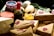 An assortment of packaged goods is displayed, including brown paper-wrapped parcels with red wax seals, a wedge of cheese with holes, a jar with a red and white checked lid, a bottle containing green peppercorns, eggs, and some leafy greens. The arrangement suggests a rustic, artisanal market setting.