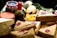 An assortment of packaged goods is displayed, including brown paper-wrapped parcels with red wax seals, a wedge of cheese with holes, a jar with a red and white checked lid, a bottle containing green peppercorns, eggs, and some leafy greens. The arrangement suggests a rustic, artisanal market setting.
