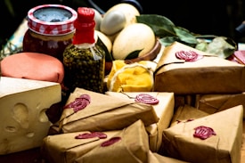 An assortment of packaged goods is displayed, including brown paper-wrapped parcels with red wax seals, a wedge of cheese with holes, a jar with a red and white checked lid, a bottle containing green peppercorns, eggs, and some leafy greens. The arrangement suggests a rustic, artisanal market setting.