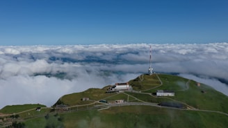 An aerial view of a lush green mountain peak with several buildings scattered across the landscape. A prominent radio or TV antenna tower stands tall on the right side. A scenic backdrop of a vast, cloud-filled sky meets the horizon, creating a serene and expansive vista.
