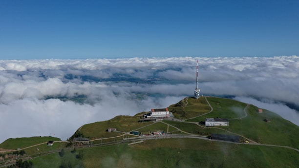 An aerial view of a lush green mountain peak with several buildings scattered across the landscape. A prominent radio or TV antenna tower stands tall on the right side. A scenic backdrop of a vast, cloud-filled sky meets the horizon, creating a serene and expansive vista.