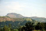 A scenic landscape featuring rolling hills covered in greenery with several wind turbines scattered across the hills. The sky is clear with a light blue hue, enhancing the serene and eco-friendly atmosphere.