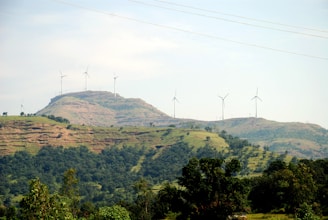 A scenic landscape featuring rolling hills covered in greenery with several wind turbines scattered across the hills. The sky is clear with a light blue hue, enhancing the serene and eco-friendly atmosphere.