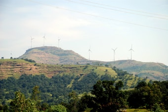 A scenic landscape featuring rolling hills covered in greenery with several wind turbines scattered across the hills. The sky is clear with a light blue hue, enhancing the serene and eco-friendly atmosphere.
